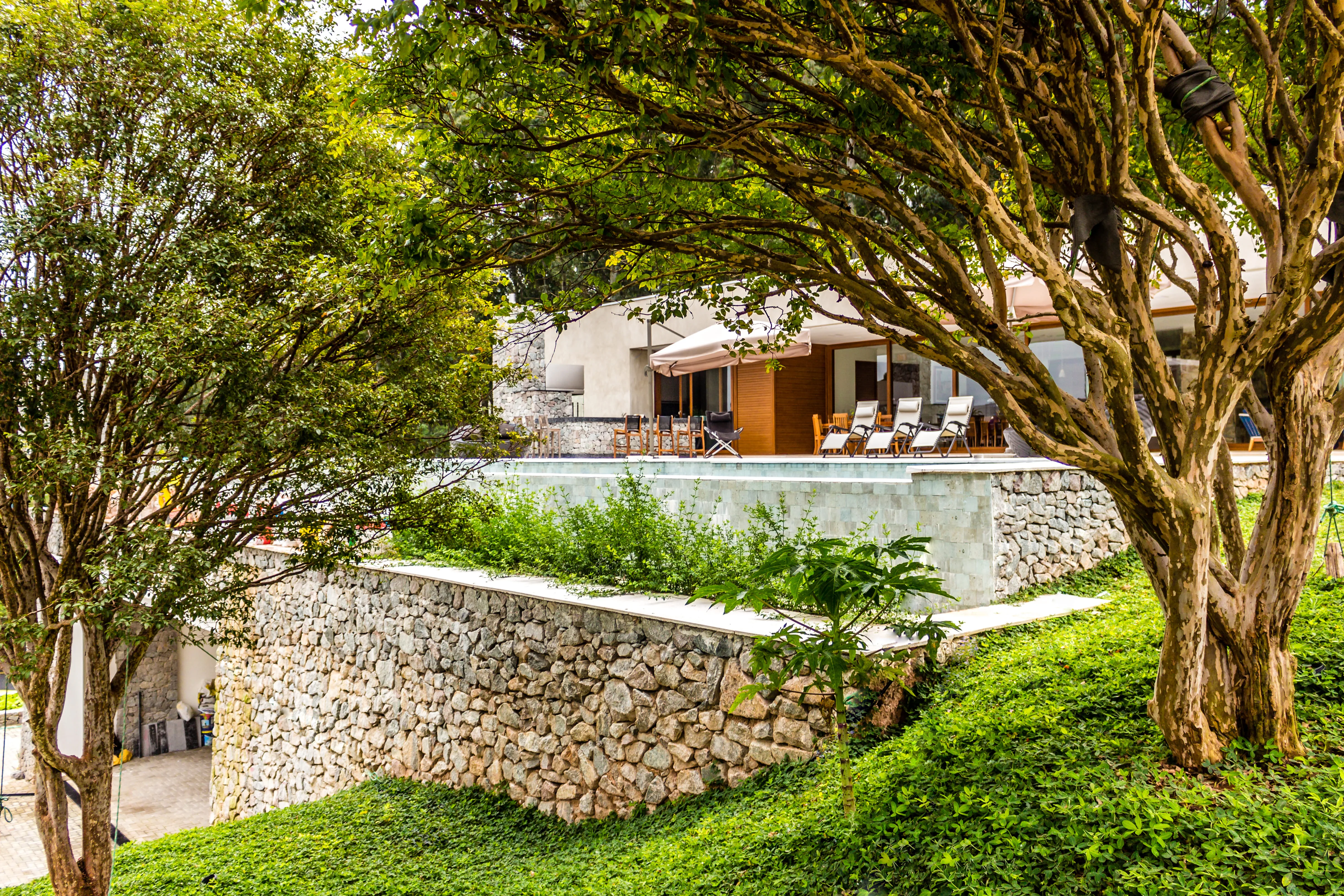 Residência contemporânea cercada por jardins e vegetação exuberante, com amplas aberturas, pedra natural, madeira e piscina. A arquitetura valoriza a conexão entre os ambientes internos, áreas externas e a natureza.